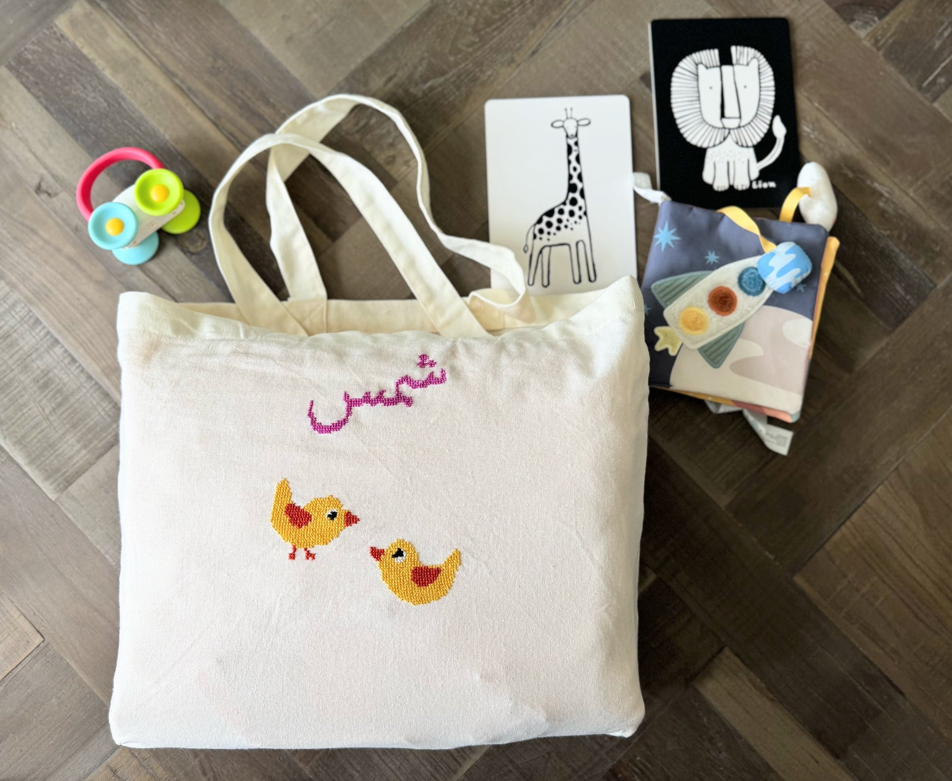 White tote bag with embroidered chicks, children's books, and toys on a wooden floor.