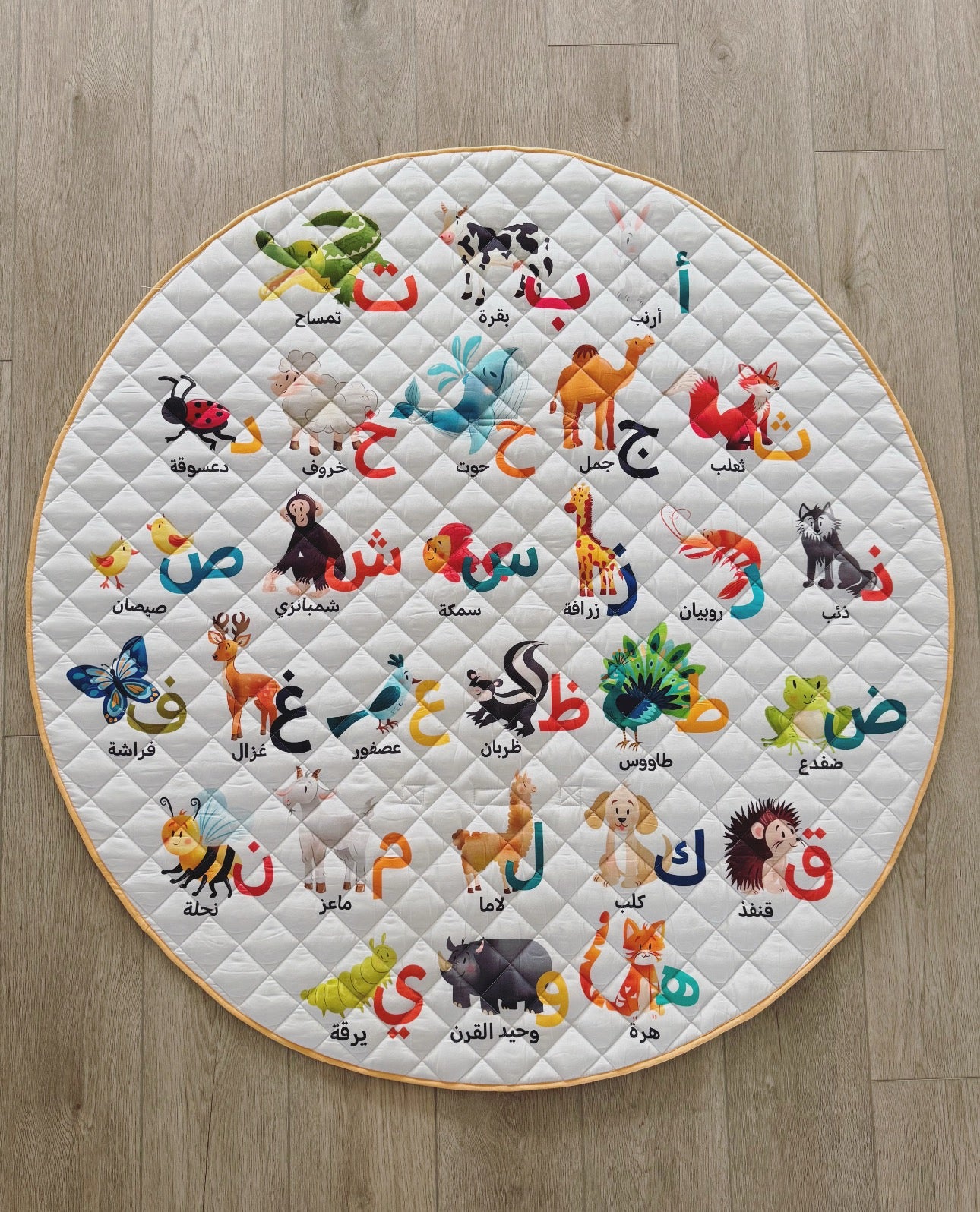 Round quilt with colorful animal illustrations and Arabic text on a wooden floor.